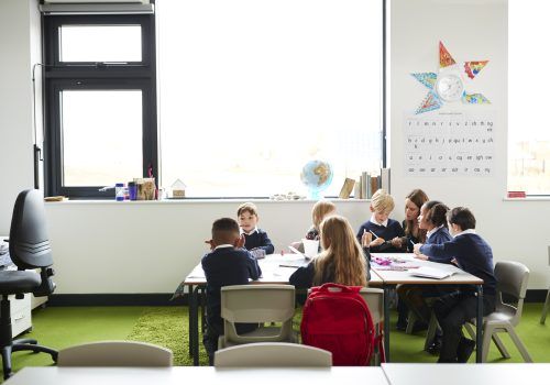A group of primary school kids sitting at table in a classroom, teacher kneeling to help them during the lesson