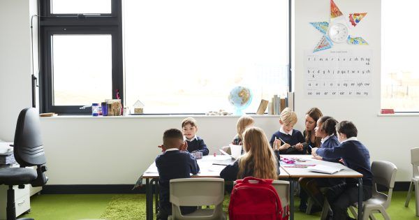 A group of primary school kids sitting at table in a classroom, teacher kneeling to help them during the lesson