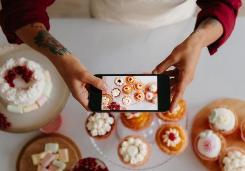Top view of female baker using smart phone while photographing sweet food laying on the table